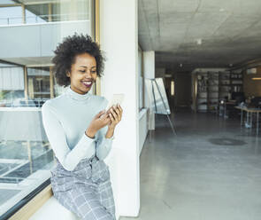 Young businesswoman using smart phone while sitting near glass window in office - UUF23507