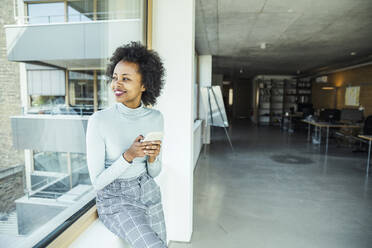 Businesswoman holding smart phone while looking away through glass window at office - UUF23508