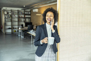 Junge Geschäftsfrau, die eine Kaffeetasse hält, während sie mit einem Smartphone im Büro telefoniert - UUF23523