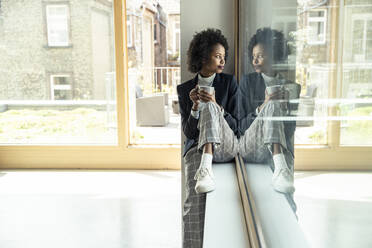 Young businesswoman with coffee cup sitting on window sill at office - UUF23525