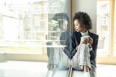 Female professional looking down while sitting on window sill at office - UUF23527