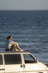 Mann auf dem Dach eines Lieferwagens sitzend mit Blick aufs Meer - OCMF02131