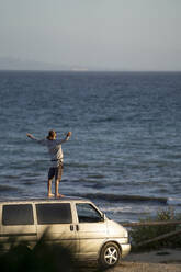 Junger Mann mit ausgestreckten Armen auf dem Dach eines Lieferwagens am Meer stehend - OCMF02133