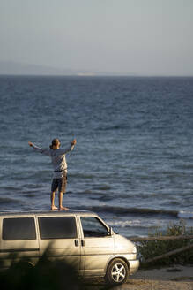 Junger Mann mit ausgestreckten Armen auf dem Dach eines Lieferwagens am Meer stehend - OCMF02133