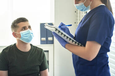 Female doctor in protective face mask taking interview of patient before vaccination at clinic - ABIF01385