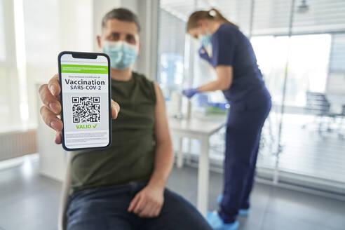 Man showing COVID-19 vaccination QR code on mobile phone while doctor in background at clinic - ABIF01395