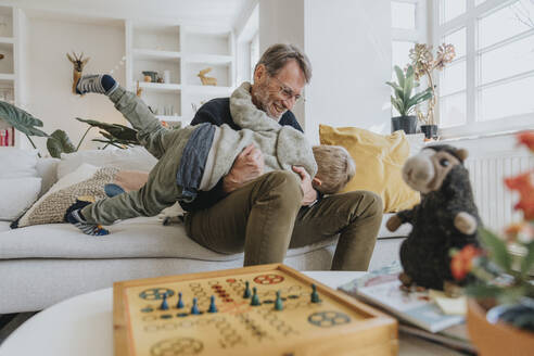 Son playing with father sitting on sofa at home - MFF07985