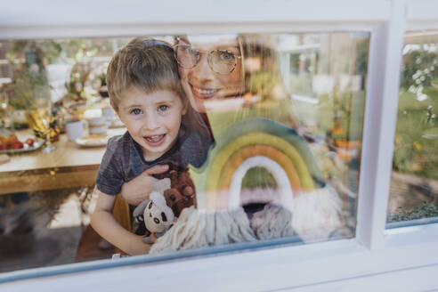 Smiling mother and son looking through window of house - MFF08007