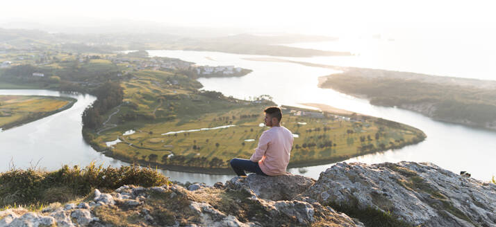 Junger Mann sitzt an einem sonnigen Tag auf einem Felsen an einer Klippe - JAQF00681