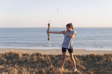 Bogenschützin zielt mit Pfeil und Bogen, während sie auf Gras am Strand steht - JRVF00992