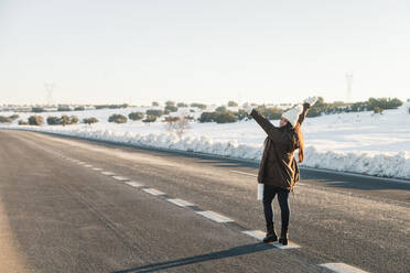 Carefree woman standing with arms raised on road during winter - MRRF01254