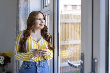 Lächelnde Frau im mittleren Erwachsenenalter, die mit ihrem Smartphone durch das Fenster schaut, während sie zu Hause steht - WPEF04735