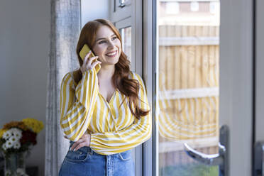 Happy redhead woman talking on smart phone looking away while standing by window - WPEF04736
