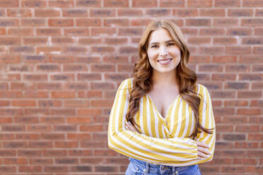 Beautiful smiling redhead woman standing with arms crossed against brick wall - WPEF04753