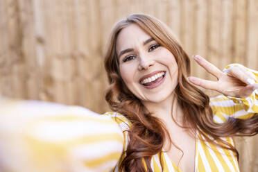 Cheerful redhead woman gesturing peace sign while standing against wooden wall - WPEF04757