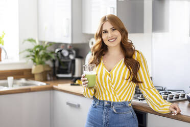 Beautiful smiling redhead woman holding healthy milkshake glass while standing in kitchen at home - WPEF04768