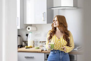 Smiling redhead woman holding milkshake glass and smart phone looking away while standing in kitchen - WPEF04770
