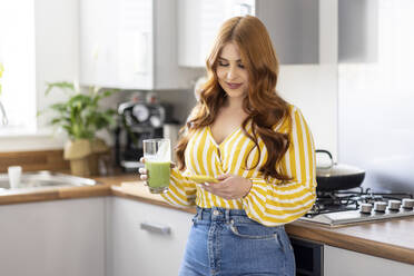 Redhead woman with milkshake glass using smart phone while standing in kitchen at home - WPEF04772