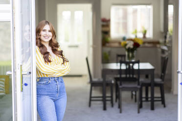 Smiling redhead woman standing with arms crossed by window - WPEF04778