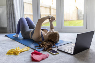 Frau auf Trainingsmatte liegend, die während des Trainings zu Hause ihre Smartwatch überprüft - WPEF04786