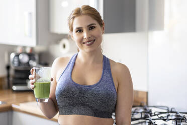 Beautiful smiling redhead woman in sports clothing holding healthy milkshake glass in kitchen - WPEF04798
