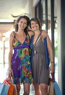 Lächelnde Freundinnen tragen Einkaufstaschen, während sie mit den Händen auf den Schultern stehen - AJOF01456