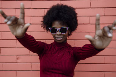 Young woman with sunglasses gesturing near brick wall - XLGF02035