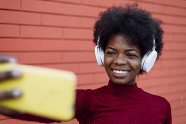 Smiling young woman with headphones taking selfie near brick wall - XLGF02038