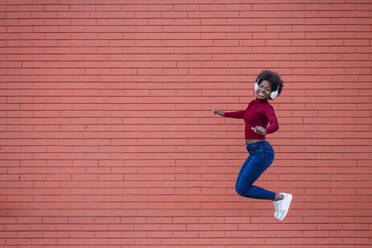 Cheerful woman with headphones jumping near brick wall - XLGF02041