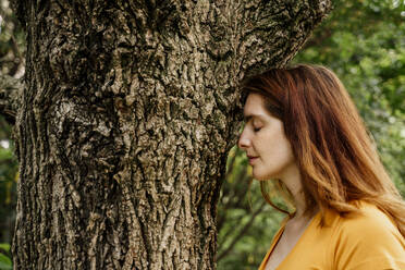 Relaxed woman with eyes closed by tree trunk in garden - AFVF08787