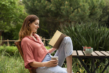 Frau mit Kaffeetasse, die auf einem Stuhl im Garten sitzend ein Buch liest - AFVF08794