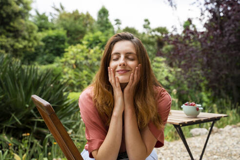 Smiling woman with eyes closed touching face while sitting on chair in garden - AFVF08797