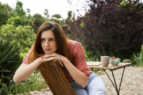 Thoughtful woman looking away while sitting on chair in garden - AFVF08799