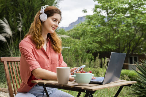 Frau mit Kopfhörern, die ein Smartphone hält, während sie einen Laptop im Garten benutzt - AFVF08801