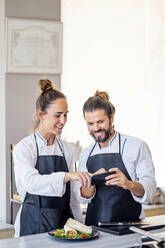 Lächelnde weibliche und männliche Köche diskutieren über das Mobiltelefon in einem Restaurant - PGF00599