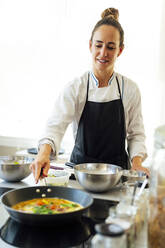 Lächelnde weibliche Köchin beim Kochen von Speisen in einer Pfanne im Restaurant - PGF00607