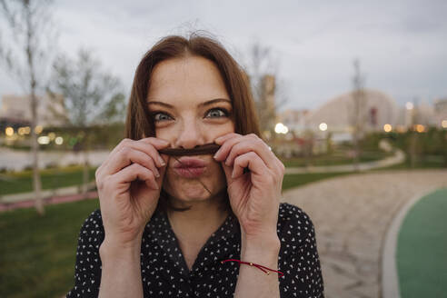 Playful mature woman making mustache with hair at park - EYAF01634