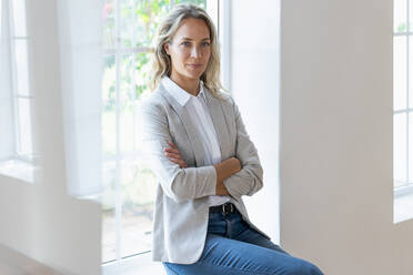 Female entrepreneur with arms crossed sitting on window sill - SBOF03915