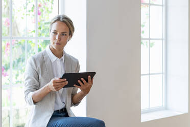 Mid adult businesswoman with digital tablet looking away while sitting on window sill - SBOF03916