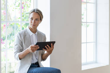 Mittlere erwachsene Geschäftsfrau mit digitalem Tablet, die auf der Fensterbank sitzt und wegschaut - SBOF03916