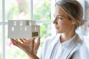 Nachdenkliche Geschäftsfrau mit Architekturmodell, die durch ein Fenster schaut - SBOF03925