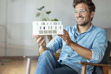 Lächelnder Geschäftsmann mit Architekturmodell auf einem Sessel im Büro sitzend - SBOF03953