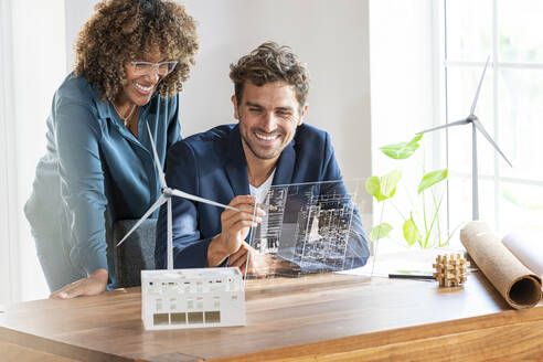 Kollegen im Büro schauen auf einen futuristischen Bildschirm mit einem Architekturplan eines nachhaltigen Hauses und Windturbinen - SBOF03971