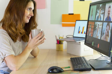 Freiberuflerin hält Tasse während eines Videogesprächs am Computer im Home Office - AFVF08804