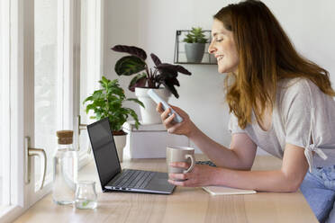 Lächelnde Frau, die ein Smartphone über einem Laptop benutzt, während sie sich im Büro zu Hause auf den Schreibtisch lehnt - AFVF08807