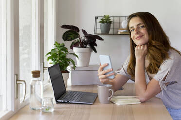 Beautiful woman with hand on chin holding smart phone at desk in home office - AFVF08808