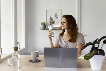 Smiling woman using smart phone at desk in home office - AFVF08813
