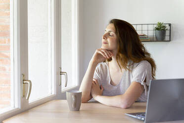 Woman with eyes closed sitting near window at home office - AFVF08816