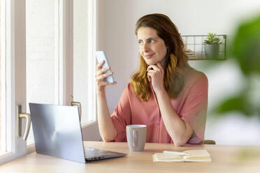 Female professional looking away while holding smart phone at home office - AFVF08817