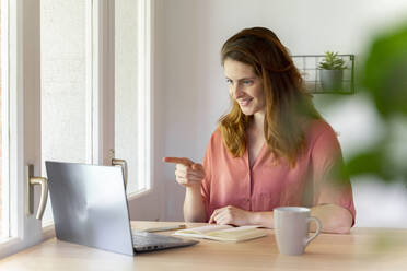Smiling businesswoman pointing during video call on laptop at home office - AFVF08819
